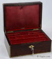 JB446: Figured rosewood and brass surround box circa 1830. A box veneered in figured rosewood box strengthened and accented with brass. The top central plaque bears initials in a stylized Gothic  script and a coronet,  probably of French nobility. There is also a brass inset of a lift-up aid on the front part of the lid. The box has extra support hinges on the sides which enable it to stay open at the same angle. The interior tray is original as is the silk faced envelope. Additional velvet covers protect and render the interior more useable. Working key. Enlarge Picture