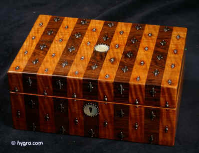 JB409: Early 19th century box veneered in alternating stripes of satinwood and rosewood and decorated with cut steel pins, faceted in the manner of precious stones. The escutcheon and central inset are in mother of pearl. Although the cut steel fine decoration is usually associated with French Palais Royale work, this is an English box possibly decorated by French highly skilled craftsmen working in England. The box has been fitted with a lift-out tray. The box has a working lock and key. Circa 1815. Enlarge Picture