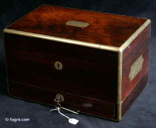 JB413: Antique George the fourth period box veneered in rosewood and edged in rounded brass for strength and contrast with the dark wood. It also has flat brass carrying handles on both sides. It has a lower jewellery drawer with a separate working lock and key which retains its original leather cover and velvet lining with a ring compartment. The top part of the box has been fitted with an extra lift-out tray. The box has two working locks and keys. Circa 1830. Enlarge Picture