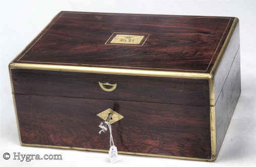 JB446: Figured rosewood and brass surround box circa 1830. A box veneered in figured rosewood box strengthened and accented with brass. The top central plaque bears initials in a stylized Gothic  script and a coronet,  probably of French nobility. There is also a brass inset of a lift-up aid on the front part of the lid. The box has extra support hinges on the sides which enable it to stay open at the same angle. The interior tray is original as is the silk faced envelope. Additional velvet covers protect and render the interior more useable. Working key. Enlarge Picture