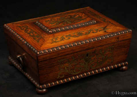 JB511: Antique box in the sarcophagus form. The box is veneered in figured rosewood and is inlaid in brass depicting stylized flora.  The fluid design of stylised flora is exceptionally fine. This is a spectacular box which encapsulates the best of the Regency era. The box stands on turned rosewood feet and has turned rosewood drop ring handles. The centre panel of the top is framed with gadrooning as is the pediment adding the architectural impact. The box has a lift out tray which has been relined . Circa1825. Enlarge Picture