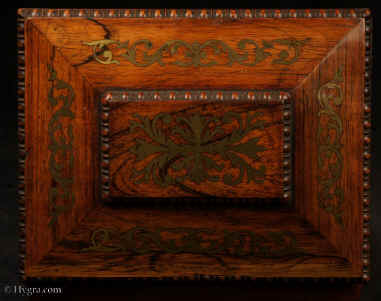 JB511: Antique box in the sarcophagus form. The box is veneered in figured rosewood and is inlaid in brass depicting stylized flora.  The fluid design of stylised flora is exceptionally fine. This is a spectacular box which encapsulates the best of the Regency era. The box stands on turned rosewood feet and has turned rosewood drop ring handles. The centre panel of the top is framed with gadrooning as is the pediment adding the architectural impact. The box has a lift out tray which has been relined . Circa1825. Enlarge Picture