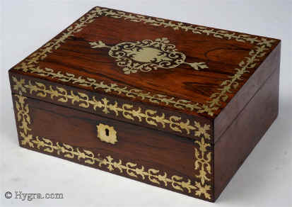 SB455: Rosewood and brass inlaid box in high Regency style circa 1815.  A box veneered in beautifully figured rosewood and inlaid with brass. The design of the inlay is of highly stylized flora, suggesting neoclassical designs hinting at palmette and (on the top) acanthus motifs. The juxtaposition of dark wood with bold brass inlay was popular in the early part of the 19th century. The wood and the bright brass were mutually enhancing. The Prince Regent (later George IV) commissioned such work for his Royal palaces. This technique, which perfected control of cutting and inlaying, required time and skill and it was very expensive at the time. It is no wonder that such work was popular at a time when excess was rife and style was given supreme social importance. The tray is beautifully made in yew wood. This is most unusual. The velvet covers are original. There are two early 19th century pin cushions which fit snugly in purposely cut-out circles. These pincushions are encased in turned and carved bone baskets. Such work is known as Dieppe work as it was mostly made by French refugees. Unfortunately the fragility of the baskets has caused them to suffer some damage, but they are quite lovely all the same. Working lock and key.
               Enlarge Picture