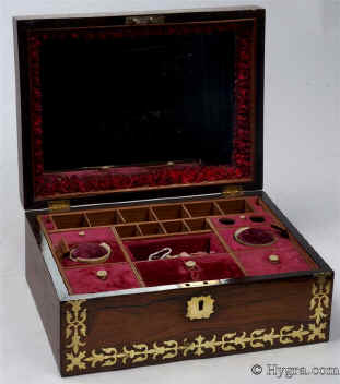 SB455: Rosewood and brass inlaid box in high Regency style circa 1815.  A box veneered in beautifully figured rosewood and inlaid with brass. The design of the inlay is of highly stylized flora, suggesting neoclassical designs hinting at palmette and (on the top) acanthus motifs. The juxtaposition of dark wood with bold brass inlay was popular in the early part of the 19th century. The wood and the bright brass were mutually enhancing. The Prince Regent (later George IV) commissioned such work for his Royal palaces. This technique, which perfected control of cutting and inlaying, required time and skill and it was very expensive at the time. It is no wonder that such work was popular at a time when excess was rife and style was given supreme social importance. The tray is beautifully made in yew wood. This is most unusual. The velvet covers are original. There are two early 19th century pin cushions which fit snugly in purposely cut-out circles. These pincushions are encased in turned and carved bone baskets. Such work is known as Dieppe work as it was mostly made by French refugees. Unfortunately the fragility of the baskets has caused them to suffer some damage, but they are quite lovely all the same. Working lock and key.
               Enlarge Picture