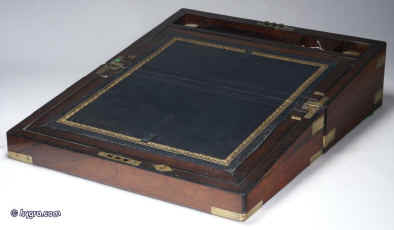 WB167: Figured rosewood writing box with brass corners and straps escutcheon and plate opening to an embossed leather (replacement) writing surface and compartments for writing implements and paper, and having two secret drawers concealed behind a sprung panel. The box has two hand blown replacement reproduction inkwells. Circa 1840 Enlarge Picture