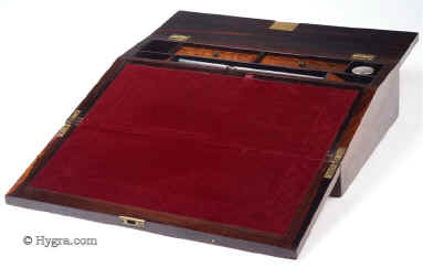 WB441: Writing slope in rosewood and satinwood with mother of pearl inlay circa 1835. This is a writing slope of exceptionally high quality. The outside is veneered in saw cut rosewood inlaid with mother of pearl. The inlay is executed with precision in two different ways: The central and corner parts have the floral patterns outlined in rosewood with the mother of pearl surrounding them; the main encircling part is made out of mother of pearl inlaid into the rosewood. On examining the rosewood patterns closely, one can see fine lines separating the leaves, a sure sign that the box retains its original finish. The design is both elegant and robust, straddling the time between the neoclassical and the naturalistic period. The patterns are symmetrical and rendered with control of line and space. This shows the neoclassical influence. There are also neoclassical motifs such as the anthemion, acanthus leaves and palmettes. However the flowers, although symmetrically arranged and stylised, have more than a hint to naturalistic aspirations and the neoclassical motifs are integrated within the whole composition. The garland, although controlled and defined within a perfect oval, loops and curls in fluid movement like a living trellis. Inside the box has a pen tray and lift up lids. The underside of the lids has at some stage been stained. There is one original inkwell with a silver plated screw on top. The writing surface is covered in subtly embossed velvet, which we think is a later replacement, but very close to what a box of this period cud have had.  The interior of the box continues the high standard of quality and it is veneered in satinwood. Small crack/repair on the top part of the veneer, perceptible when the box is closely examined.  Working lock and key. Enlarge Picture