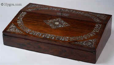 WB441: Writing slope in rosewood and satinwood with mother of pearl inlay circa 1835. This is a writing slope of exceptionally high quality. The outside is veneered in saw cut rosewood inlaid with mother of pearl. The inlay is executed with precision in two different ways: The central and corner parts have the floral patterns outlined in rosewood with the mother of pearl surrounding them; the main encircling part is made out of mother of pearl inlaid into the rosewood. On examining the rosewood patterns closely, one can see fine lines separating the leaves, a sure sign that the box retains its original finish. The design is both elegant and robust, straddling the time between the neoclassical and the naturalistic period. The patterns are symmetrical and rendered with control of line and space. This shows the neoclassical influence. There are also neoclassical motifs such as the anthemion, acanthus leaves and palmettes. However the flowers, although symmetrically arranged and stylised, have more than a hint to naturalistic aspirations and the neoclassical motifs are integrated within the whole composition. The garland, although controlled and defined within a perfect oval, loops and curls in fluid movement like a living trellis. Inside the box has a pen tray and lift up lids. The underside of the lids has at some stage been stained. There is one original inkwell with a silver plated screw on top. The writing surface is covered in subtly embossed velvet, which we think is a later replacement, but very close to what a box of this period cud have had.  The interior of the box continues the high standard of quality and it is veneered in satinwood. Small crack/repair on the top part of the veneer, perceptible when the box is closely examined.  Working lock and key.Enlarge Picture